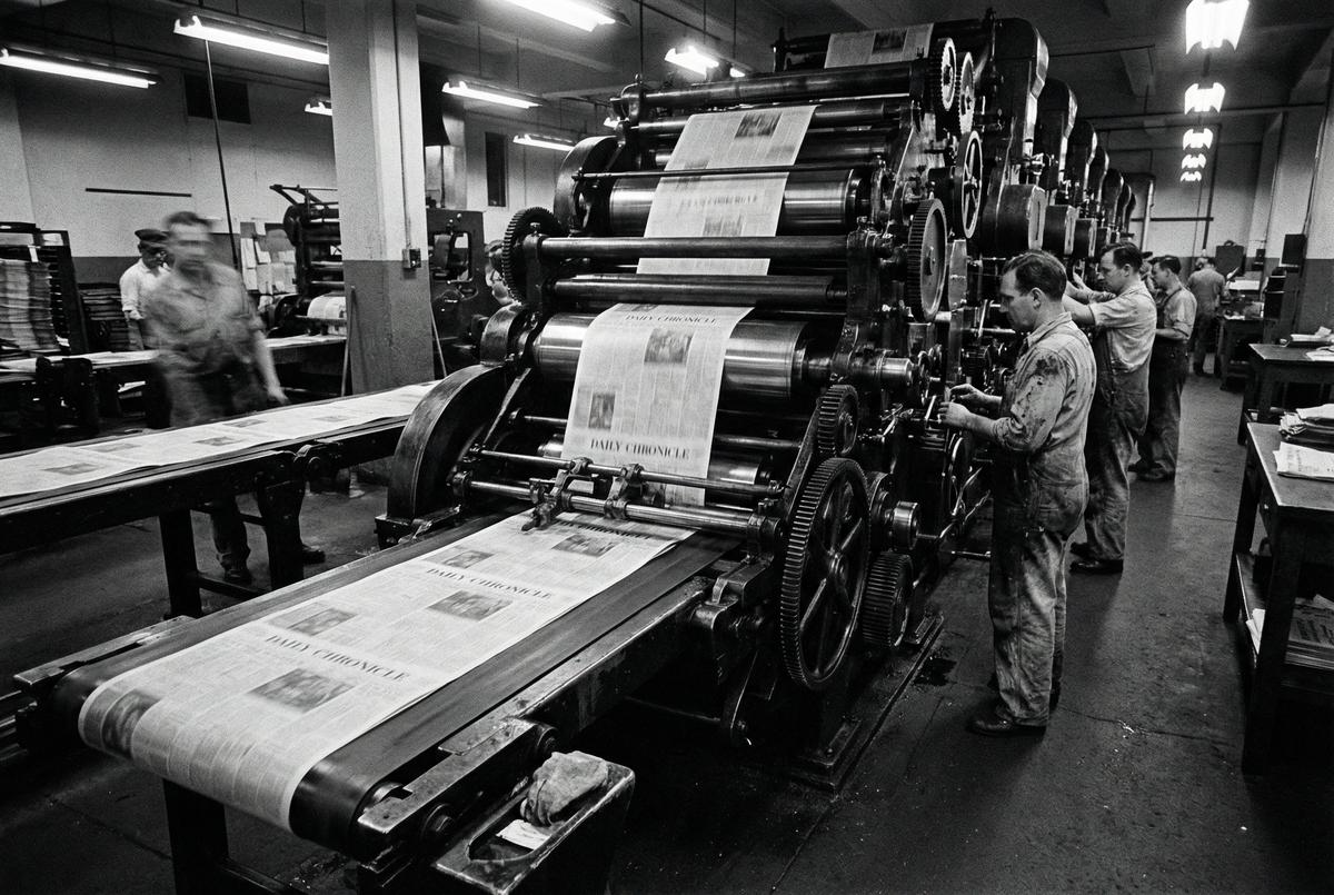 Workers operating a large newspaper printing press, ink-heavy rolls turning, pages emerging at speed.
